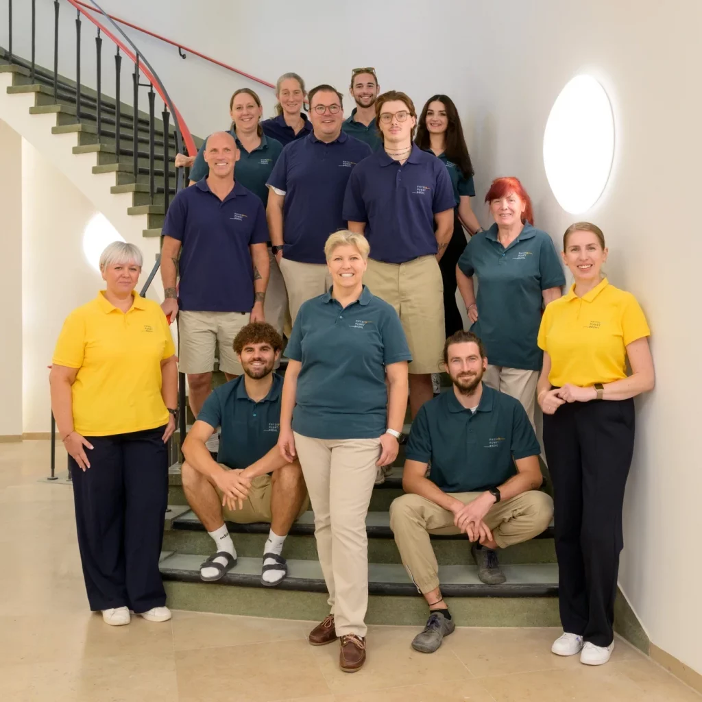 Das gesamte Team von Physiopunkt Brühl steht gemeinsam auf einer Treppe in der Praxis und lächelt in die Kamera – Physiotherapeutinnen und Physiotherapeuten in moderner Arbeitskleidung