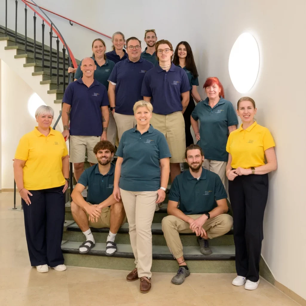 Das gesamte Team von Physiopunkt Brühl steht gemeinsam auf einer Treppe in der Praxis und lächelt in die Kamera – Physiotherapeutinnen und Physiotherapeuten in moderner Arbeitskleidung.